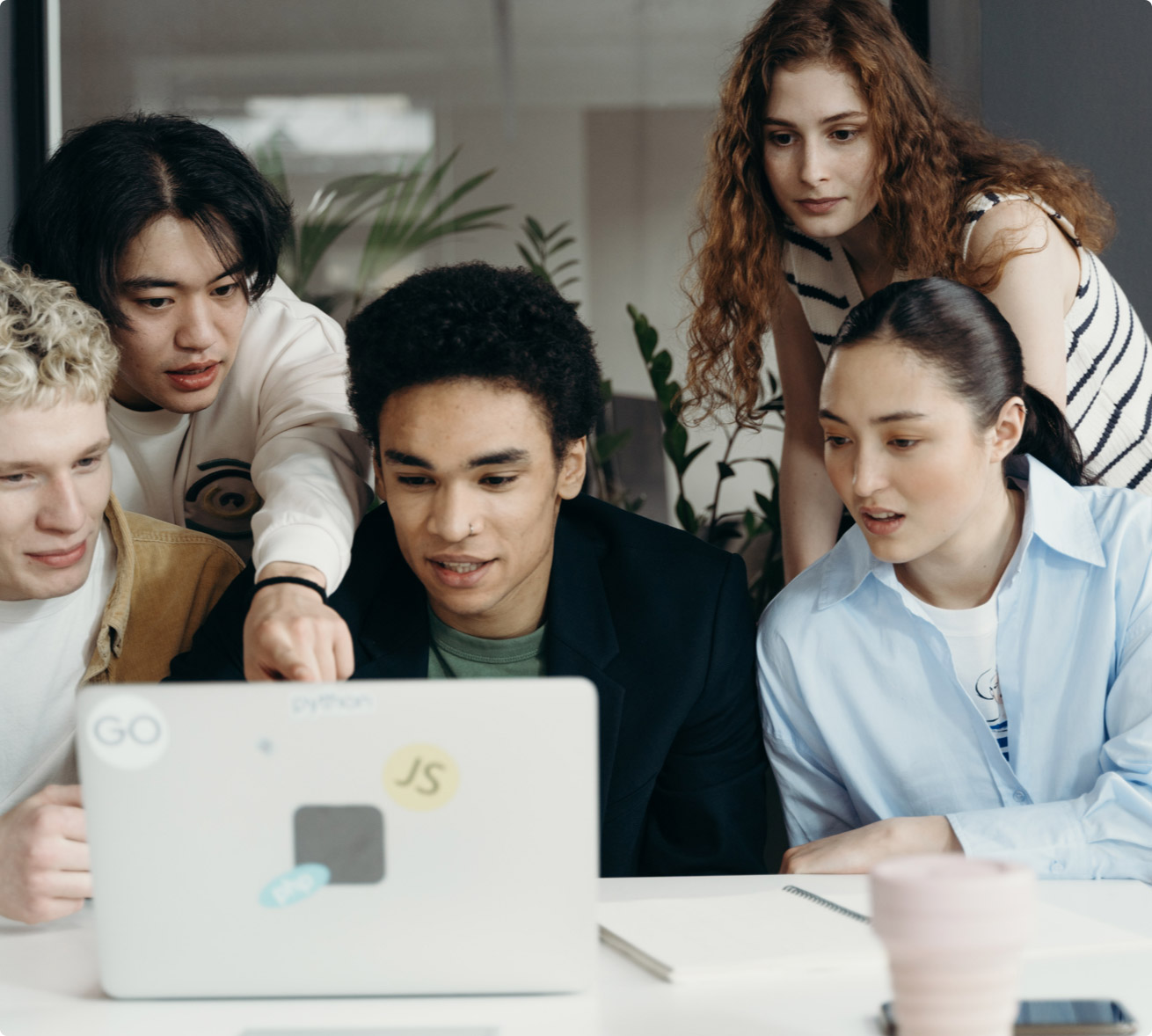 Three diverse software developers laughing and collaborating on a project around a laptop and a whiteboard filled with diagrams in a bright, modern co-working space.